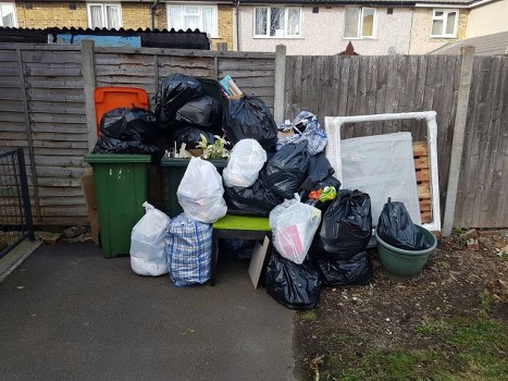 A collection of black and white rubbish bags filled with waste are piled on and around a small green outdoor bench situated on a paved driveway, adjacent to a wooden fence. Some of the bags appear to contain household waste, while others are tied and placed in a disorderly manner. Behind the pile, there is a large orange wheeled bin and a green wheeled bin, both positioned against the wooden fence. An old, disused window frame or picture frame with a white, worn finish leans against the fence on the right side of the scene. To the right of the rubbish bags, a small round green plastic container is visible on the ground. The background includes the lower parts of residential buildings with visible windows, indicative of a typical Milton Keynes residential area, suggesting this is an outdoor rubbish collection or disposal site. The scene is lit by natural daylight, with the overall setting emphasizing the need for waste removal services such as those provided by Commercial Waste Milton Keynes, which handles rubbish clearance in local communities.