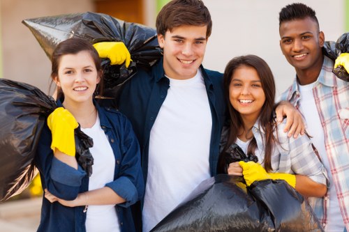 Four young volunteers, two women and two men, stand outdoors on a sunny day, each holding large black trash bags filled with rubbish, prepared for collection. The volunteers are dressed casually, with some wearing yellow gloves and smiling at the camera. They are positioned closely together, with a suburban or park environment in the background, featuring blurred greenery and a paved area. The scene suggests a community clean-up activity, potentially organized by Commercial Waste Milton Keynes, a professional rubbish removal service operating in the Milton Keynes area. The black rubbish bags and the volunteers’ positive attitude highlight their efforts to clear waste and improve the local environment, fitting within the context of a waste management initiative. The lighting is natural, emphasizing the casual, outdoor setting and the clean, tidy appearance of the rubbish bags meant for disposal or collection.