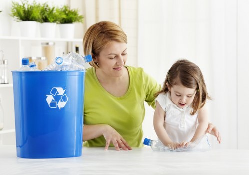 A woman and a young girl are engaged in recycling activities in a bright, well-lit kitchen with white shelving units and potted plants in the background. The woman, dressed in a lime-green top, is seated at a light-colored kitchen table, guiding the girl in sorting or preparing plastic bottles for disposal. The girl, wearing a white dress, is focused on handling a clear plastic bottle, with her hands placed on the table. To the left, a large blue recycling bin with a white recycling symbol is positioned on the table; it contains several crumpled plastic bottles and other recyclable plastics. The scene highlights responsible waste management practices with natural daylight illuminating the scene, emphasizing the importance of recycling and waste sorting within a domestic setting. This visual aligns with the services of Commercial Waste Milton Keynes who support effective rubbish removal for local households and businesses in the MK region, fostering environmentally conscious waste disposal methods.
