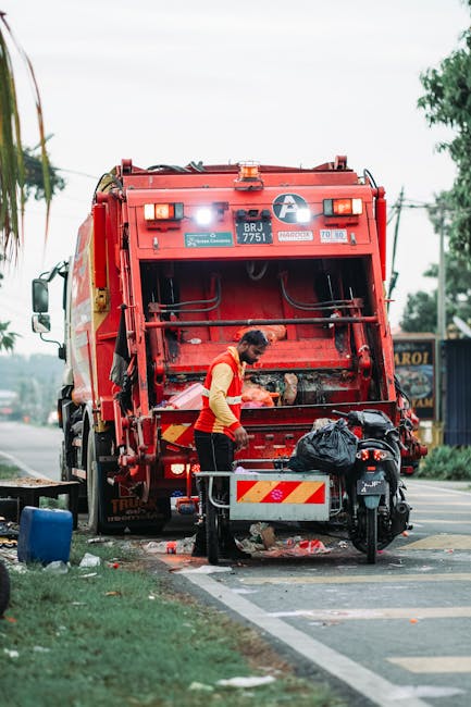 A worker in a high-visibility yellow vest and dark clothing is operating a commercial rubbish collection vehicle on a city street during dusk. The worker's back is facing the camera, and they are usin